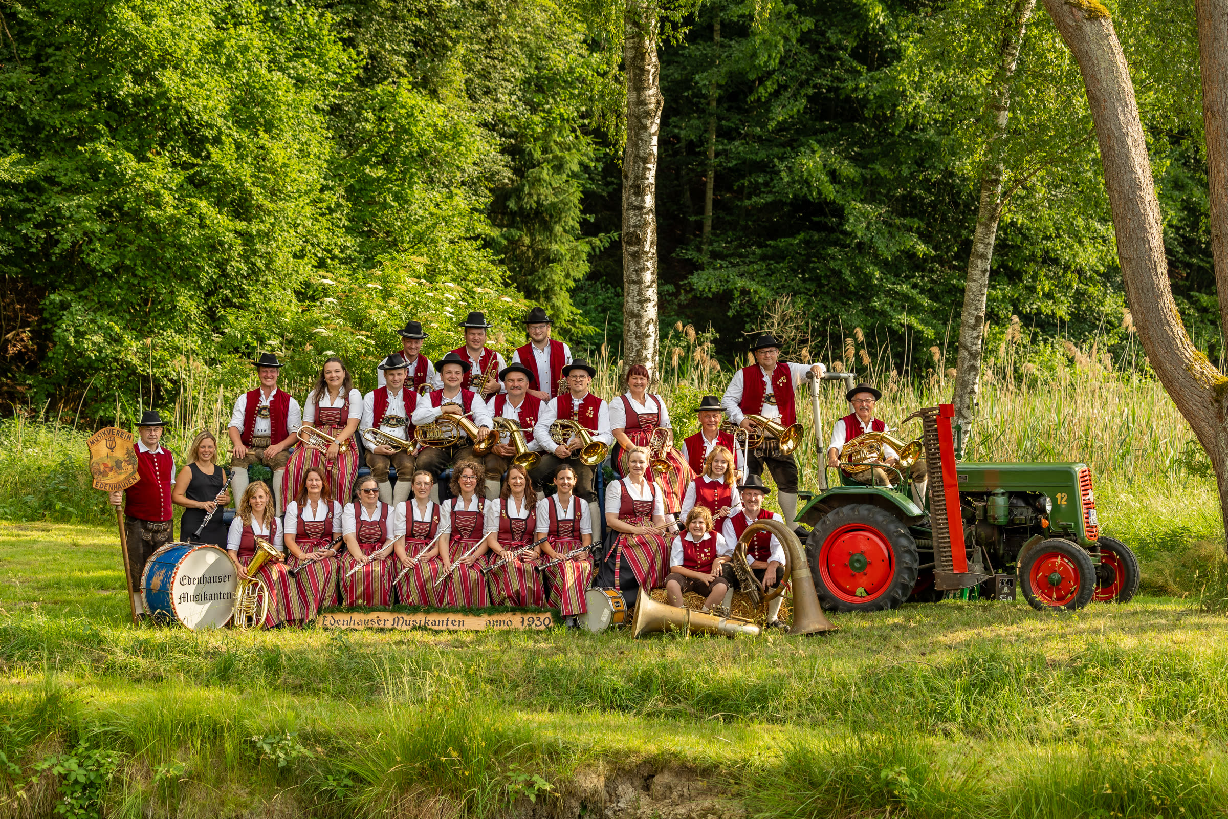 Gruppenbild 2026: die ganze Kapelle des Musikverein Edenhausen in Tracht am Waldrand, mit Bulldog und dem Schild „Edenhauser Musikanten anno 1930“